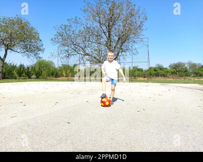 Sremska Mitrovica, Serbien. 6. Juni 2020. Ein Junge spielt Ball auf dem Spielplatz. Asphaltsportplatz. Ein Kind in einem weißen T-Shirt. Kleinkind mit blondem Haar, 7 Jahre alt. Laufen, Treten und Übungen Stockfoto