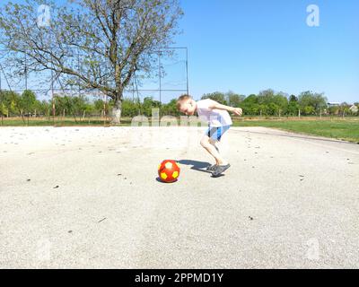 Sremska Mitrovica, Serbien. 6. Juni 2020. Ein Junge spielt Ball auf dem Spielplatz. Asphaltsportplatz. Ein Kind in einem weißen T-Shirt. Kleinkind mit blondem Haar, 7 Jahre alt. Laufen, Treten und Übungen Stockfoto