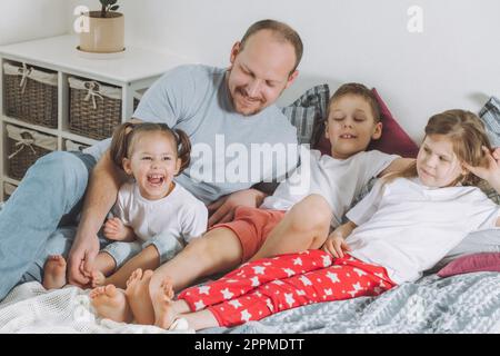 Vater spielt mit drei Kindern im Bett. Dad kitzelt Kinderfüße. Eine Familie von Daddy, zwei Mädchen und ein Junge Stockfoto