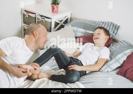 Vater spielt mit seinem Sohn 7-10 im Bett. Dad kitzelt Kinderfüße. Familie, Spaß haben Stockfoto