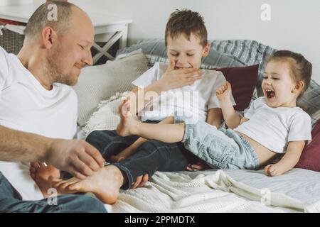 Vater spielt mit zwei Kindern im Bett. Dad kitzelt Kinderfüße. Familie von Daddy, Mädchen und Jungen Stockfoto