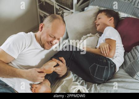Vater spielt mit seinem Sohn 7-10 im Bett. Dad kitzelt Kinderfüße. Familie, Spaß haben Stockfoto