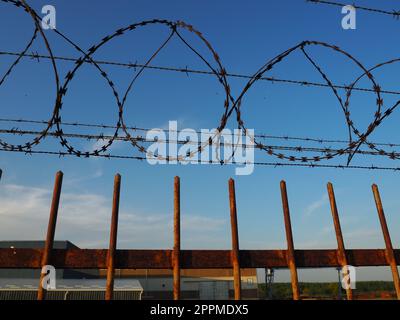 Stacheldraht im Strang und gedehnt gegen den blauen Himmel. Stacheldraht gegen Diebe und Vandalen. Ein unüberwindbares Hindernis. Rostige Drähte, Reihen, Spiralen und Spulen von Stacheldraht. Stockfoto