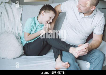 Vater spielt mit seinem Sohn 7-10 auf der Couch. Dad kitzelt Kinderfüße. Familie, Spaß haben Stockfoto