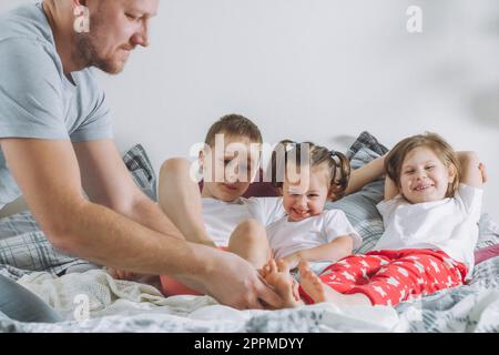 Vater spielt mit drei Kindern im Bett. Dad kitzelt Kinderfüße. Eine Familie von Daddy, zwei Mädchen und ein Junge Stockfoto