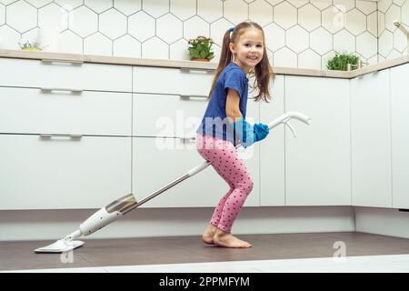 Glückliches kleines Mädchen, das auf einem modernen Wischmopp sitzt, um den Boden zu waschen, mit abnehmbarer Bürste neben dem Küchenset. Hausreinigungskonzept. Stockfoto