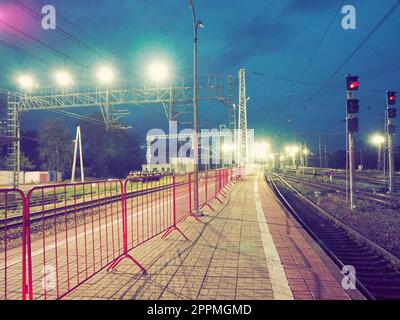 Chudovo, Russland 6. August 2021 Bahnhof bei Nacht, Plattform mit Plattform. An der Ampel leuchtet die rote Ampel. Mehrere Weitstrahler beleuchten den Weg. Der Metallzaun ist rot gestrichen Stockfoto