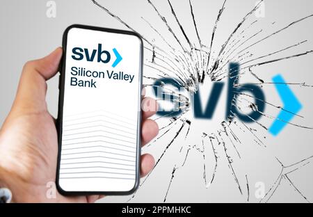Hand hält ein Telefon mit Silicone Valley Bank-Website auf dem Bildschirm mit zerbrochenem Glas und verschwommenem SVB-Logo im Hintergrund Stockfoto
