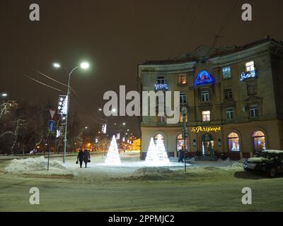 Petrozavodsk, Karelien, Russland, 01. Januar 2021. Karl Marx Avenue, Café Parisianka am Abend im Winter. Schneeverwehungen, Straßenbeleuchtung und Dekorationen. Die Leute gehen die Straße entlang. Stockfoto