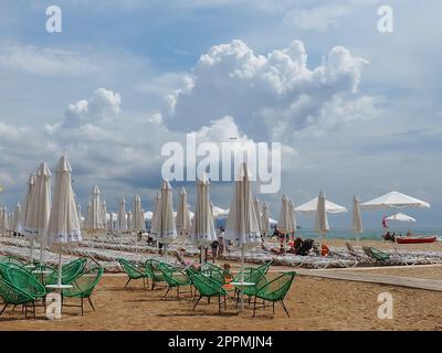 Anapa, Russland, 15. August 2021 Sonnenliegen und Sonnenschirme am Sandstrand nach dem Regen. Strandsaison. Strandcafe mit Korbstühlen. Touristen, die Leute laufen lassen. Eine Sturmwarnung. Kumuluswolken Stockfoto