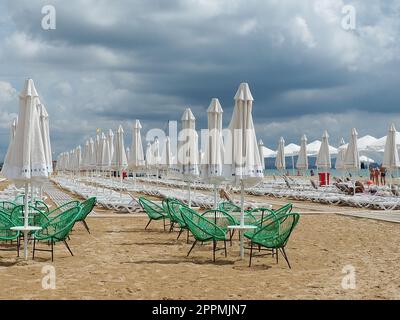 Anapa, Russland, 15. August 2021 Sonnenliegen und Sonnenschirme am Sandstrand nach dem Regen. Strandsaison. Strandcafe mit Korbstühlen. Touristen, die Leute laufen lassen. Eine Sturmwarnung. Kumuluswolken Stockfoto