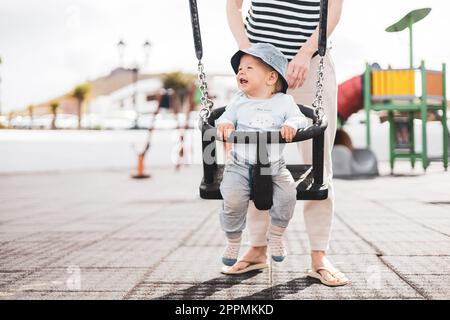 Mutter schubst ihr kleines Kind auf einer Schaukel auf dem Spielplatz im Freien. Stockfoto