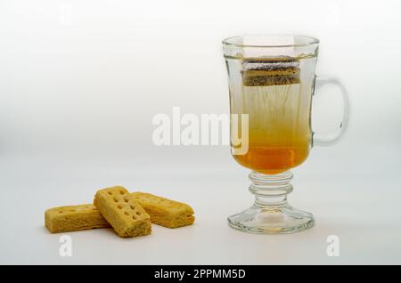 Heißer Tee, der in einem klaren Becher mit Strähnen aus Teeessenz serviert wird und das Wasser mit dem wunderbaren Geschmack der Blätter und Short-Brote erfüllt Stockfoto