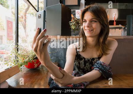 Junge transsexuelle Latina-Frau argentinischer Herkunft lächelt, macht ein Selfie in einem Restaurant, während sie darauf wartet, serviert zu werden. Stockfoto
