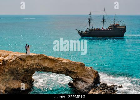 Ein paar frisch Vermählte, Braut und Bräutigam, stehen auf der felsigen Love Bridge in Zypern, nahe Ayia Napa. Vor dem Hintergrund fährt ein Schiff im Retro-Stil durch das Meer Stockfoto