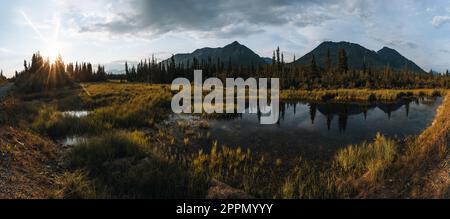 Blick auf die McCarthy Road, McCarthy Valley, Wrangell St. Elias-Nationalpark, Alaska. Wiesen und Berge mit einem See bei Sonnenuntergang. Tundra-Landschaft. Stockfoto