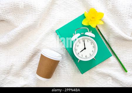 Buch, Wecker, Kaffeetasse und Blume im weißen Bett, Draufsicht. Stockfoto