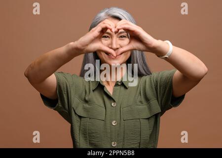 Charmante, grauhaarige Asiatin mit Herzzeichen und isolierten Handflächen auf braunem Hintergrund. Liebe und Zuneigung Stockfoto