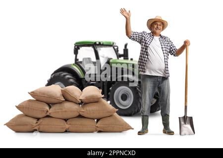Ein vollständiges Porträt eines reifen Landwirts mit einem Spaten und einem Stapel Säcke vor einem grünen Traktor, der lächelt und winkt, isoliert auf weißem Hintergrund Stockfoto