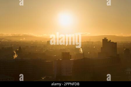 Sonnenaufgang über den Dächern von Manchester an einem Wintermorgen, mit Rauch aus den Türmen und Häusern in der Ferne, warmes Licht am Himmel über der Stadt Stockfoto