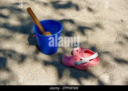 Outdoor Sandspielzeug und ein paar Sandalen im Sandkasten im Schatten eines Baumes im Sommer. Stockfoto