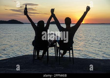 Ein Paar beobachtet den Sonnenuntergang auf den Philippinen, sitzt auf Stühlen und hebt die Arme mit einer Flasche Wein. Aufgenommen auf den 100 Inseln. Stockfoto
