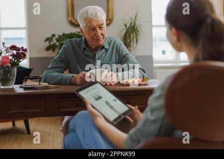 Pflegekraft, die Online-Zeitung auf einem Tablet liest, während sie sich um Senioren kümmert. Stockfoto