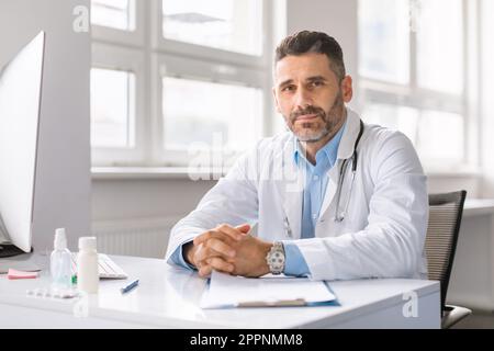Porträt eines selbstbewussten männlichen Arztes in Arbeitskleidung, der in der Klinik arbeitet, am Schreibtisch mit Computer sitzt und in die Kamera schaut Stockfoto