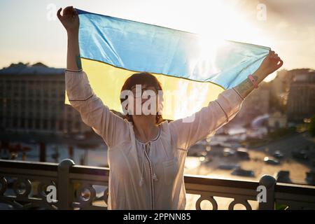 Eine bezaubernde Weiße in Silhouette in weißer Bluse und Brille hält die Flagge der Ukraine, winkt im Wind mit Blick auf die Stadt und dem Leuchteffekt der Linse. Patriotische Frau mit ukrainischer Flagge Stockfoto