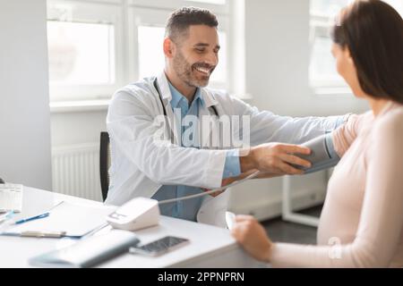 Professionelle Versorgung während der Schwangerschaft. Männlicher Arzt mittleren Alters, der die Herzfrequenz und den Blutdruck schwangerer Frauen überprüft Stockfoto