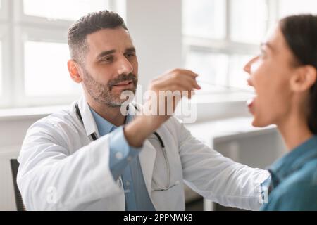 Männlicher Arzt mittleren Alters, der die Kehle einer Patientin untersucht, Therapeut, die die Kehlkopfuntersuchung der Frau durchführt Stockfoto