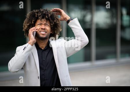 Außenporträt eines wütenden schwarzen Geschäftsmanns mittleren Alters, der mit dem Handy spricht und mit den Händen Gesten macht und draußen steht Stockfoto