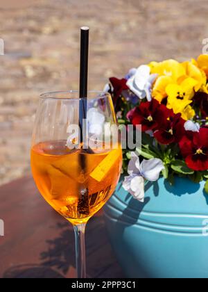 Ein erfrischendes Aperol Spritz garniert mit einer blühenden Pflanze, serviert in einem gelben Weinglas für die perfekte alkoholische Erfrischung. Stockfoto