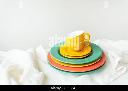 Leere gelbe Tassen und bunte Teller auf einem weißen Tisch, der mit einer Leinentischdecke bedeckt ist Stockfoto