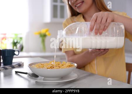 Junge Frau, die Milch aus einer Gallonflasche in den Teller gießt, mit Frühstücksflocken am weißen Marmortisch in der Küche, Nahaufnahme Stockfoto