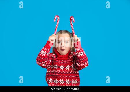 Überraschtes kleines Mädchen in gestricktem Weihnachts-Pullover mit Zuckerstangen auf blauem Hintergrund Stockfoto