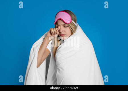 Junge Frau mit Schlafmaske, eingewickelt mit weicher Decke auf blauem Hintergrund Stockfoto