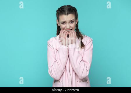 Portrait eines nervösen gestressten Teenagers mit Zöpfen in rosa Jacke, das in tiefer Depression ist, Panikattacken fühlt, ihre Fingernägel beißt. Innenstudio-Aufnahme isoliert auf grünem Hintergrund. Stockfoto