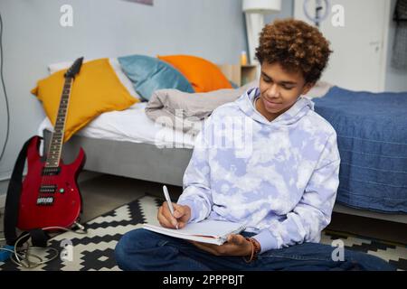 Jugendlicher Student in Freizeitkleidung, der sich Notizen in einem Copybook macht, während er auf dem Boden mit einem Doppelbett sitzt und die Hausaufgabe vorbereitet Stockfoto