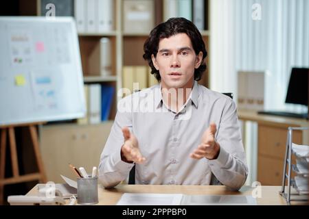 Junger, selbstbewusster, männlicher Geschäftscoach, der etwas auf dem Seminar erklärt, während er vor Publikum oder einer Gruppe von Kollegen am Schreibtisch sitzt Stockfoto