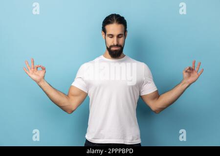 Porträt eines ruhigen, entspannten Mannes mit Bart, der ein weißes T-Shirt trägt, Yoga macht, atmet, die Augen geschlossen hält und sich nach der Arbeit beruhigt. Innenstudio, isoliert auf blauem Hintergrund. Stockfoto