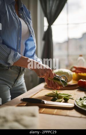 Eine Frau, die eine Gurke zum Mittagessen schält. Hausfrau, die eine Gemüsehaut mit einem Schälwerkzeug abzieht Stockfoto