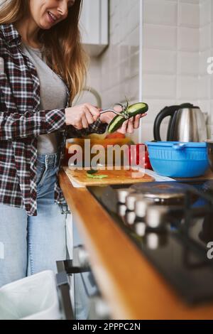 Fröhliche Erwachsene Frau, die eine Gurkenhaut mit einem Schälwerkzeug abzieht. Weibliche Person, die zu Hause frischen Salat kocht Stockfoto