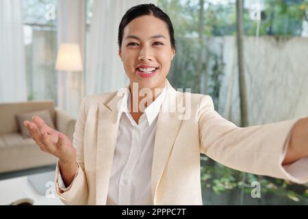 Junge, fröhliche Bloggerin oder Unternehmerin, die mit einem Online-Publikum zu Hause spricht und dabei auf die Smartphone-Kamera schaut Stockfoto