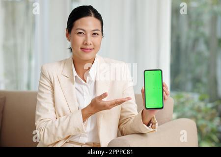 Fröhliche junge brünette Geschäftsfrau, die neue mobile App präsentiert, während sie ein Smartphone mit grünem Bildschirm hält und in die Kamera schaut Stockfoto