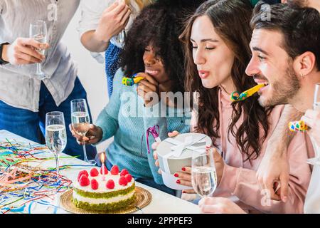 Geburtstagsparty mit Freunden. Ein Mädchen bläst Kerzen auf Kuchen aus und hält ihr Geschenk, bevor es ausgepackt wird Stockfoto