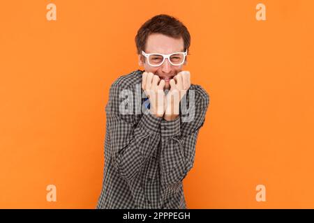 Ein ängstlicher verwirrter Mann, der Nerd beißt Fingernägel, ist unsicher und nervös, fühlt sich unwohl, trägt ein Hemd mit blauer Fliege und einer weißen Brille. Innenstudio, isoliert auf orangefarbenem Hintergrund. Stockfoto