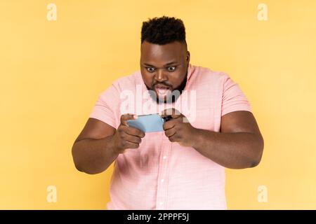 Porträt eines erstaunten Mannes, der ein pinkfarbenes Hemd im Stehen trägt, ein Smartphone benutzt und mit aufgeregtem Gesicht und großen Augen ein mobiles Spiel spielt. Innenstudio-Aufnahme isoliert auf gelbem Hintergrund. Stockfoto