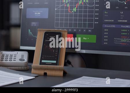 Nahaufnahme eines Smartphones mit Finanzdiagrammen auf dem Bildschirm, der auf einem Tisch steht und Computer im Büro hat Stockfoto