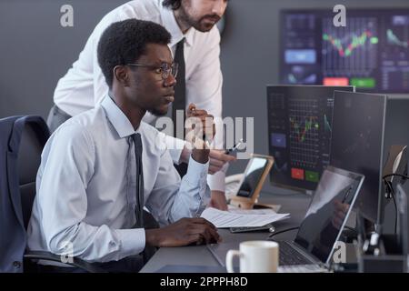 Junge Makler, die auf hohe Indikatoren warten, auf Computerbildschirme schauen, während sie im Maklerbüro am Tisch sitzen Stockfoto